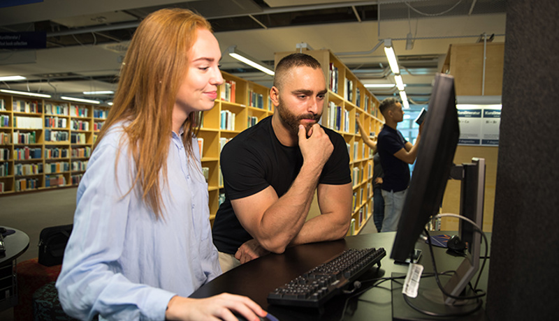 Två personer framför en dator i ett bibliotek.